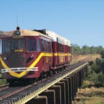 The Gulflander RM93 train crosses a heritage bridge surrounded by outback vegetation under a clear blue sky.