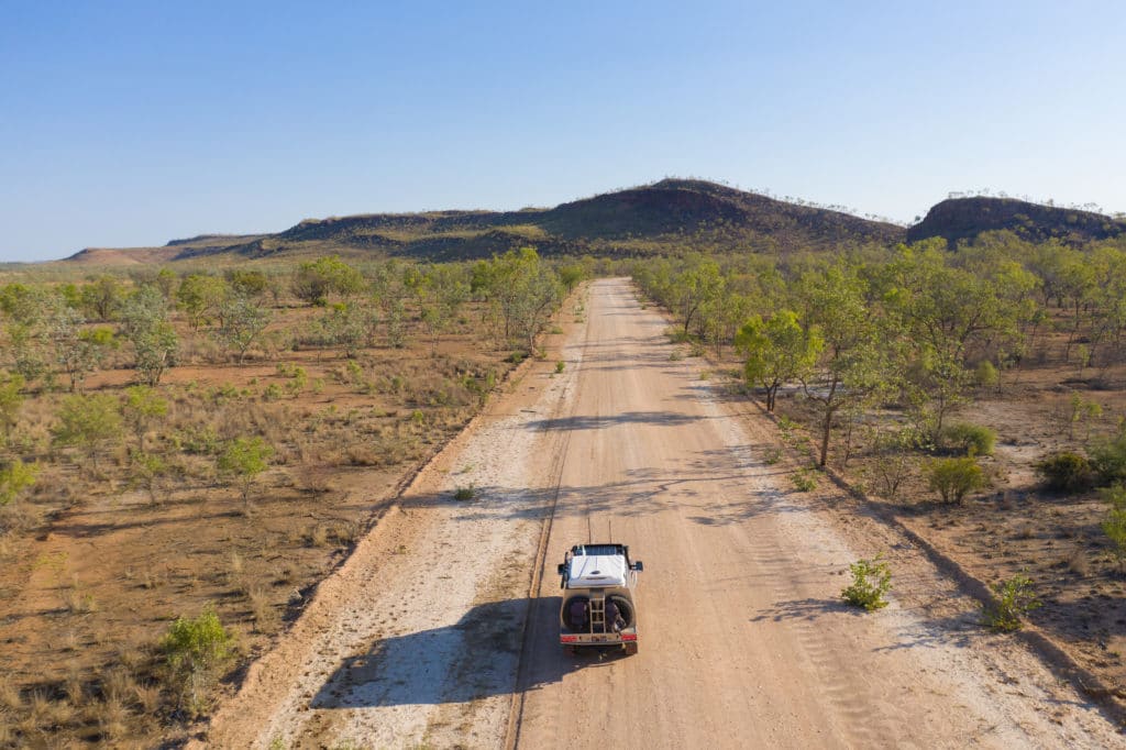 A 4WD vehicle travels down a dusty outback road surrounded by bushland and low hills in the Gulf Savannah region.