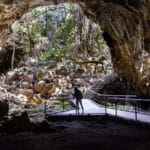 Person standing at the entrance of an ancient lava tube surrounded by boulders and rainforest at Undara Volcanic National Park, explored on a Gulf Savannah Tours adventure.