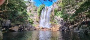 A wide waterfall cascading into a crystal-clear rock pool surrounded by rainforest trees and boulders, with people wading at the base.