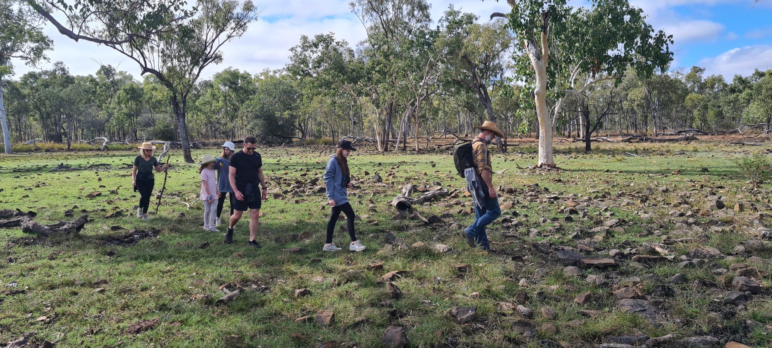 A small group of adults and children walking through grassy and rocky terrain in the Gulf Savannah, surrounded by gum trees.
